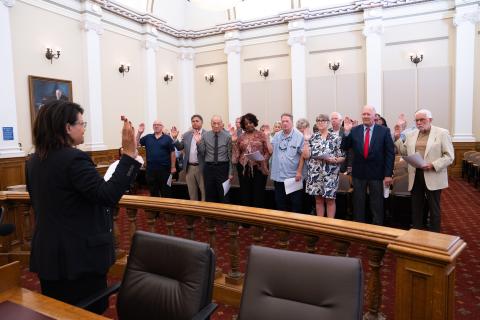 Presiding Judge Jacqueline C. Jackson administers the oath to a group of jurors with raised hands in a courtroom with white columns and wood-paneled jury box railings.